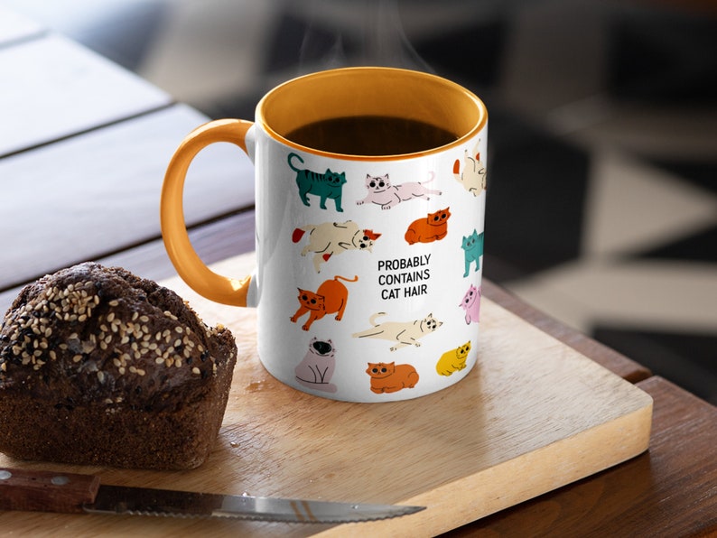 May include: White ceramic mug with an orange rim and handle. The mug features a pattern of colourful cats and the text "Probably Contains Cat Hair".