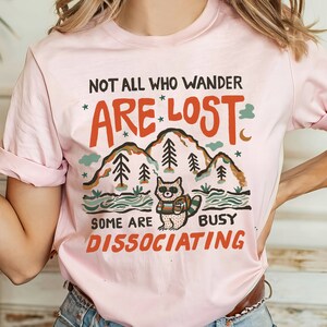 May include: Pink t-shirt with a graphic of a raccoon wearing a backpack and hiking in the mountains. The text reads "Not all who wander are lost. Some are busy dissociating."