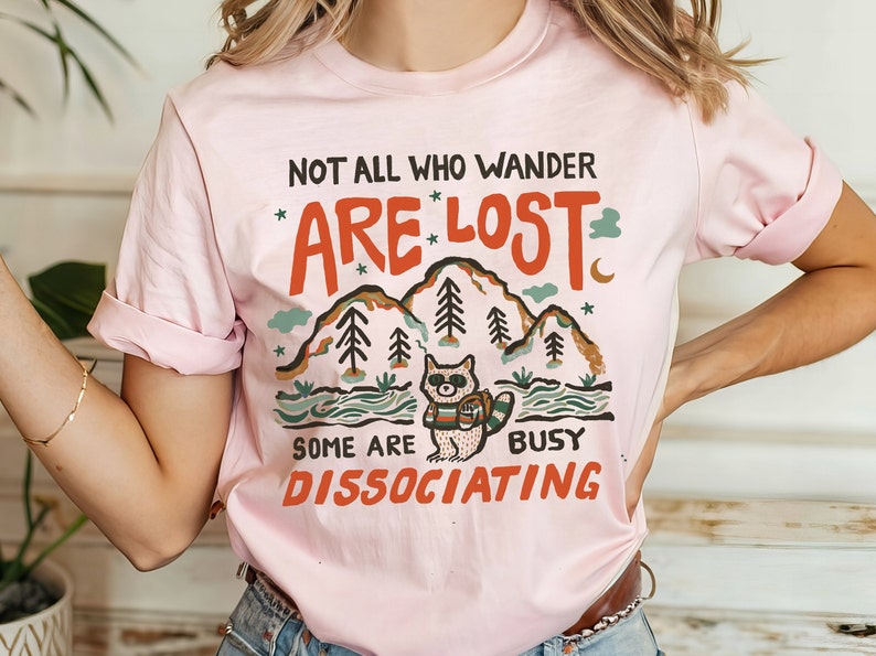 May include: Pink t-shirt with a graphic of a raccoon wearing a backpack and hiking in the mountains. The text reads "Not all who wander are lost. Some are busy dissociating."