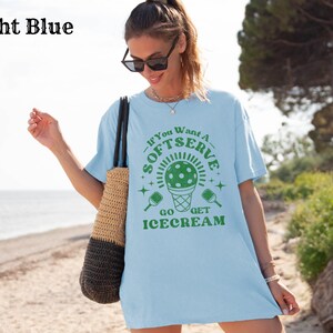 以下が含まれることがあります： 薄い青色のTシャツに、緑色のピクルボールとアイスクリームコーンのグラフィックが描かれています。Tシャツのテキストは「ソフトサーブが欲しいなら、アイスクリームを買いに行きましょう。」です
