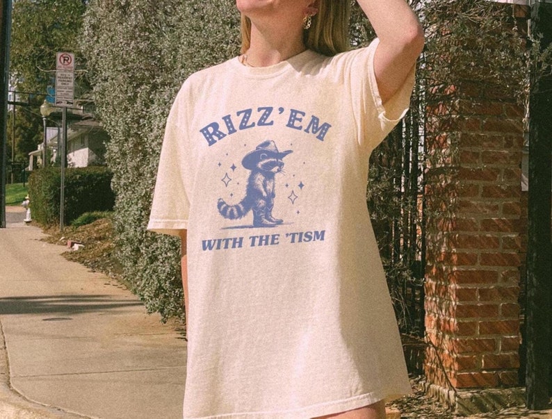 Peut inclure: Un t-shirt beige clair avec un graphique bleu d'un raton laveur portant un chapeau de cowboy et le texte "Rizz 'Em With The Tism".