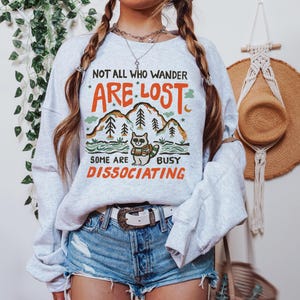 May include: A gray sweatshirt with a graphic print of a raccoon in a forest scene. The text on the sweatshirt reads "Not all who wander are lost, some are busy disassociating."