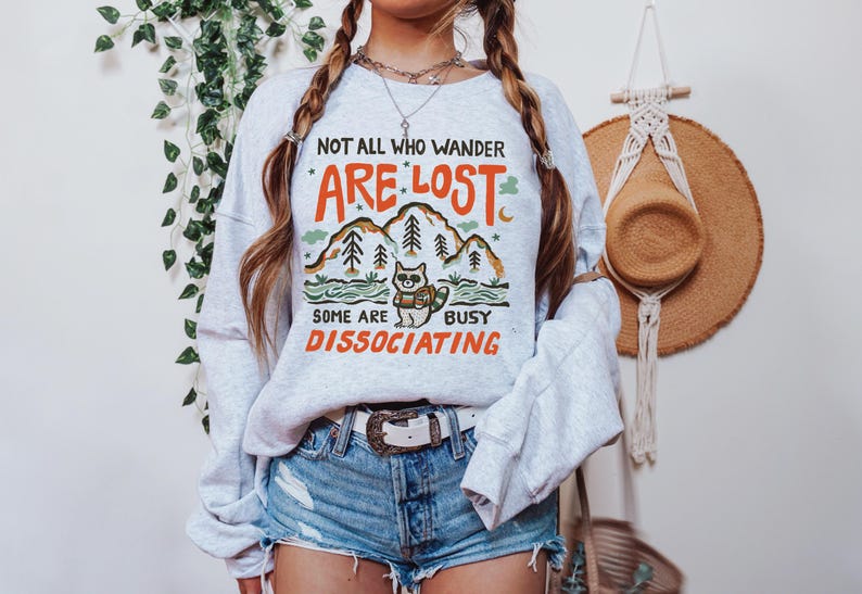 May include: A gray sweatshirt with a graphic print of a raccoon in a forest scene. The text on the sweatshirt reads "Not all who wander are lost, some are busy disassociating."