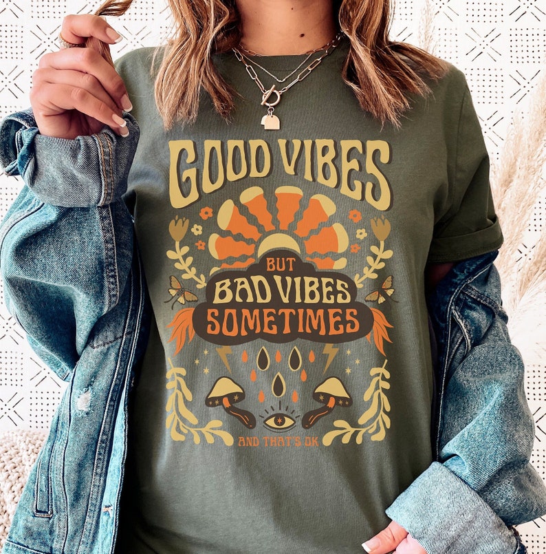 May include: Green t-shirt with a retro graphic design that says "Good Vibes But Bad Vibes Sometimes And That's Ok". The graphic features a sun, flowers, mushrooms, and a third eye.