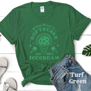 以下が含まれることがあります： 白い文字で「If You Want A Softserve Go Get Icecream」と書かれた緑色のTシャツ。デザインには、ピクルボールが入った緑と白のアイスクリームコーンも含まれています。Tシャツは、白い木製の表面に、白いサンダル、赤いサングラス、緑の葉のキーホルダーと一緒に置かれています。