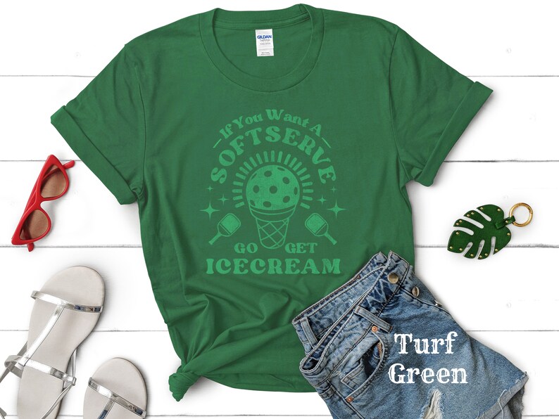 以下が含まれることがあります： 白い文字で「If You Want A Softserve Go Get Icecream」と書かれた緑色のTシャツ。デザインには、ピクルボールが入った緑と白のアイスクリームコーンも含まれています。Tシャツは、白い木製の表面に、白いサンダル、赤いサングラス、緑の葉のキーホルダーと一緒に置かれています。