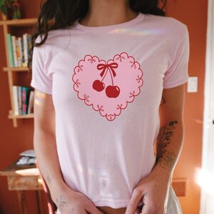 Peut inclure: T-shirt rose avec un motif en forme de c&oelig;ur rouge repr&eacute;sentant deux cerises rouges avec un n&oelig;ud rouge sur le dessus.