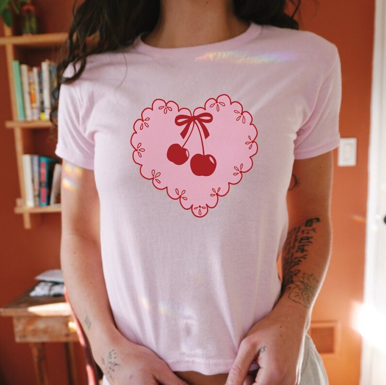 Peut inclure: T-shirt rose avec un motif en forme de c&oelig;ur rouge repr&eacute;sentant deux cerises rouges avec un n&oelig;ud rouge sur le dessus.