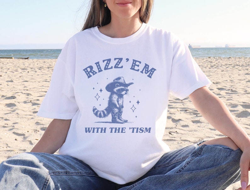 Peut inclure: T-shirt blanc avec un graphique bleu d'un raton laveur portant un chapeau de cowboy et le texte "Rizz 'Em With The 'Tism".
