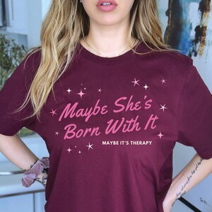 Puede incluir: Una camiseta color burdeos con el texto "Maybe She's Born With It" en letras rosas y el texto "Maybe It's Therapy" en letras rosas m&aacute;s peque&ntilde;as. La camiseta est&aacute; decorada con peque&ntilde;as estrellas rosas.