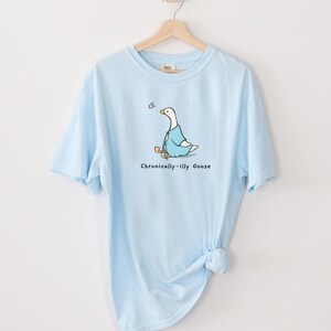 May include: A light blue t-shirt with a cartoon illustration of a goose wearing a blue jacket and holding a stick. The text "Chronically-illy Goose" is printed below the goose.