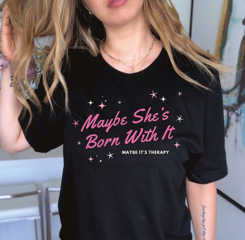 Puede incluir: Camiseta negra con texto rosa que dice "Maybe She's Born With It" y "Maybe It's Therapy" con estrellas rosas alrededor del texto.