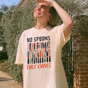 May include: A cream coloured t-shirt with a graphic print of knives and flowers. The text "NO SPOONS ONLY KNIVES" is printed on the shirt.