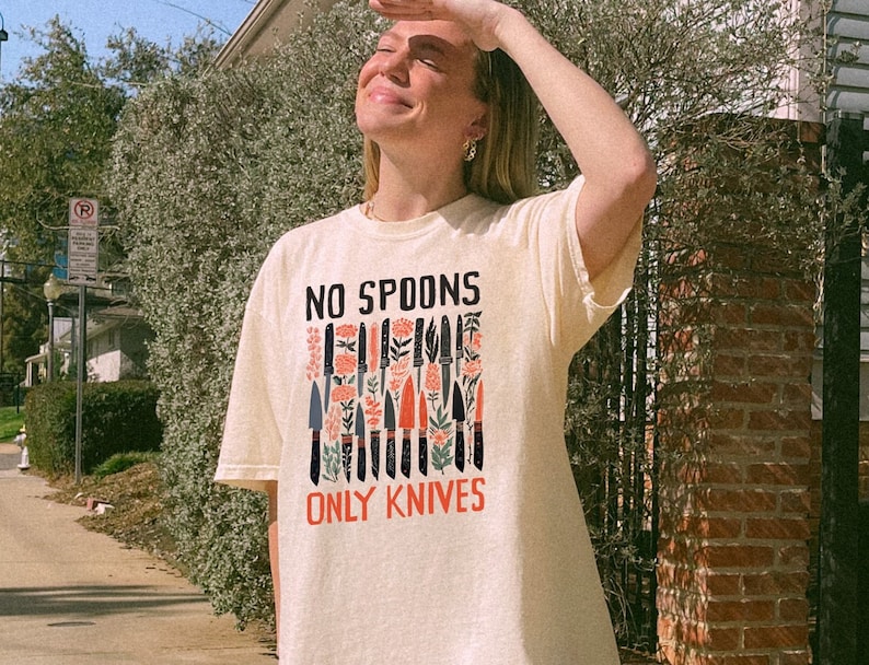 May include: A cream coloured t-shirt with a graphic print of knives and flowers. The text "NO SPOONS ONLY KNIVES" is printed on the shirt.