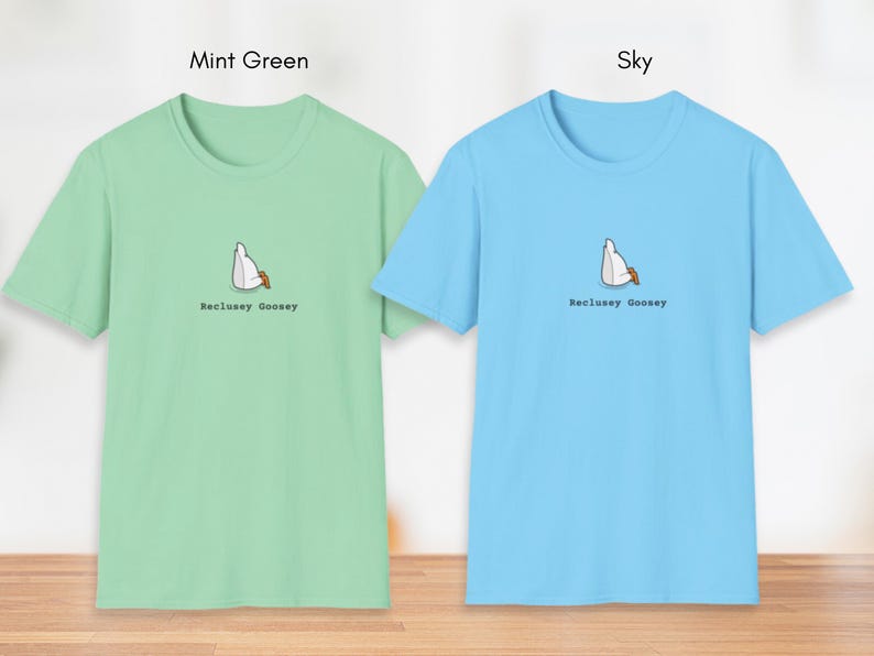 May include: Two t-shirts with a graphic of a goose sitting in a chair. The t-shirt on the left is mint green and the t-shirt on the right is sky blue. Both t-shirts have the text "Reclusey Goosey" printed on them.
