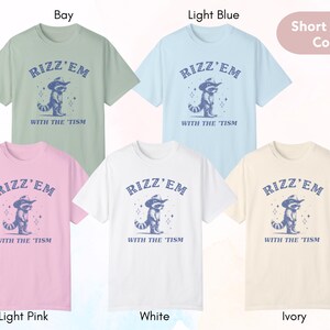 Peut inclure: Six t-shirts &agrave; manches courtes de diff&eacute;rentes couleurs, chacune repr&eacute;sentant un raton laveur portant un chapeau de cowboy et le texte "Rizz'em with the 'tism". Les couleurs sont baie, bleu clair, rose clair, blanc et ivoire.