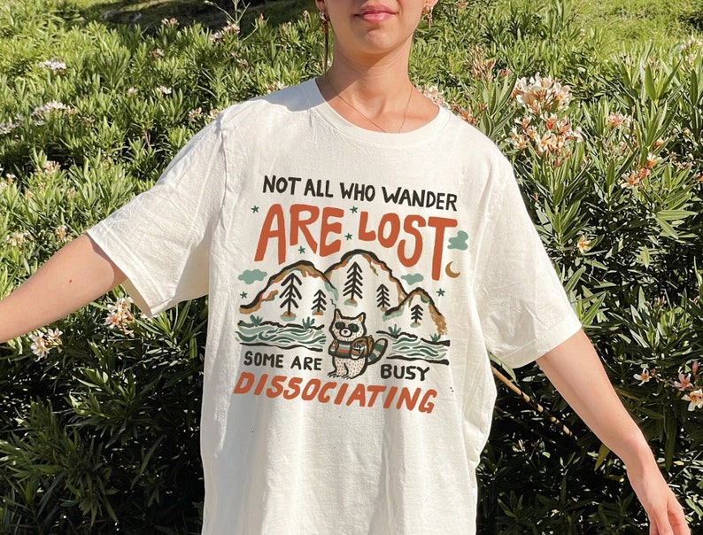 May include: White t-shirt with a graphic of a raccoon in a forest with the text "Not all who wander are lost. Some are busy dissociating."