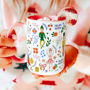 Puede incluir: Una taza de cerámica rosa con fondo blanco y un diseño navideño colorido con una bailarina, un cascanueces y un árbol de Navidad. La taza está llena de chocolate caliente y cubierta de malvaviscos. Un bastón de caramelo sobresale de la taza.