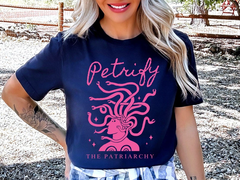K&ouml;nnte beinhalten: Ein marineblaues T-Shirt mit einer pinkfarbenen Grafik von Medusa mit Schlangen als Haaren. Der Text "Petrify The Patriarchy" ist auf das Shirt gedruckt.