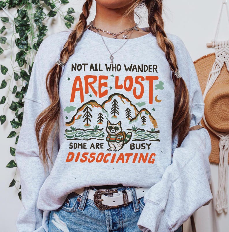 May include: A gray sweatshirt with a graphic print of a raccoon in a forest with the text "Not all who wander are lost. Some are busy dissociating."