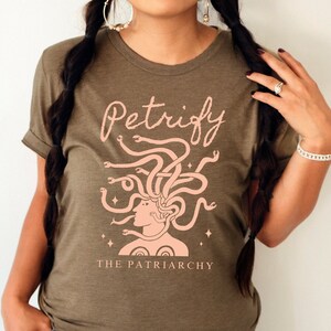 K&ouml;nnte beinhalten: Ein gr&uuml;nes T-Shirt mit einem rosafarbenen Motiv einer Frau mit Schlangen als Haaren. Der Text "Petrify the Patriarchy" ist unter dem Motiv aufgedruckt.