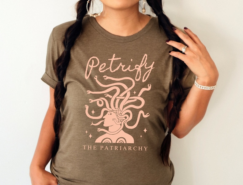 K&ouml;nnte beinhalten: Ein gr&uuml;nes T-Shirt mit einem rosafarbenen Motiv einer Frau mit Schlangen als Haaren. Der Text "Petrify the Patriarchy" ist unter dem Motiv aufgedruckt.