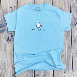 May include: A light blue t-shirt with a cartoon goose sitting on its feet and the text "Reclusey Goosey".