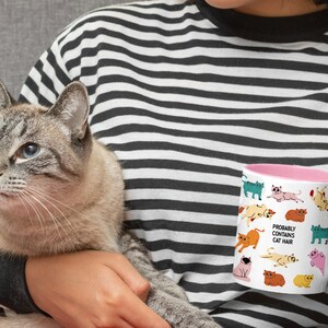 May include: A white ceramic mug with a pink rim features a whimsical design of various cats in different poses. The text "Probably Contains Cat Hair" is printed on the mug.