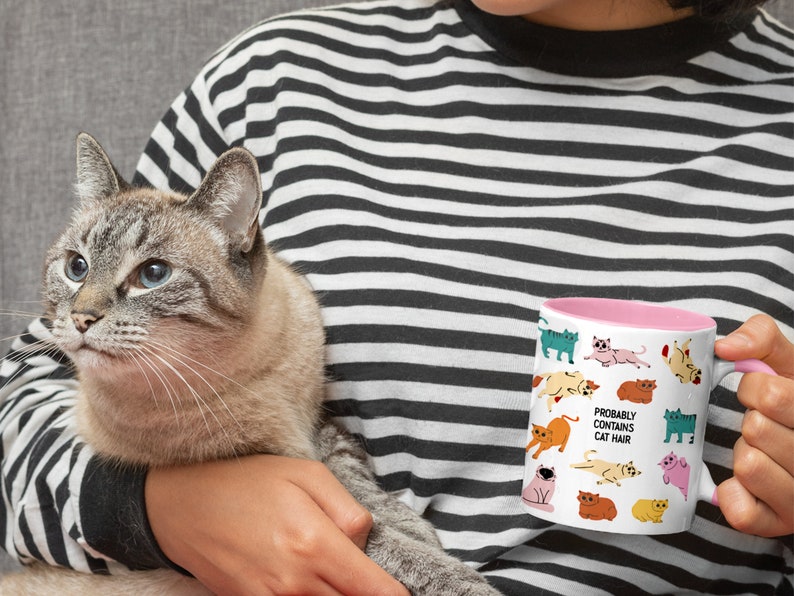 May include: A white ceramic mug with a pink rim features a whimsical design of various cats in different poses. The text "Probably Contains Cat Hair" is printed on the mug.
