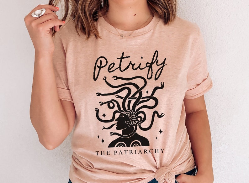 K&ouml;nnte beinhalten: Ein hellrosa T-Shirt mit einer schwarzen Grafik einer Frau mit Schlangen als Haaren. Der Text "Petrify The Patriarchy" ist unter der Grafik aufgedruckt.