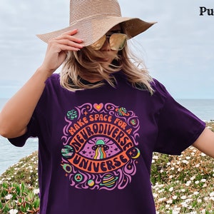 May include: A woman wearing a purple t-shirt with the text "Make Space for a Neurodiverse Universe" in a colourful, psychedelic font. The shirt features a graphic of an eye with a UFO flying over it. The woman is standing in front of a body of water and a field of flowers.