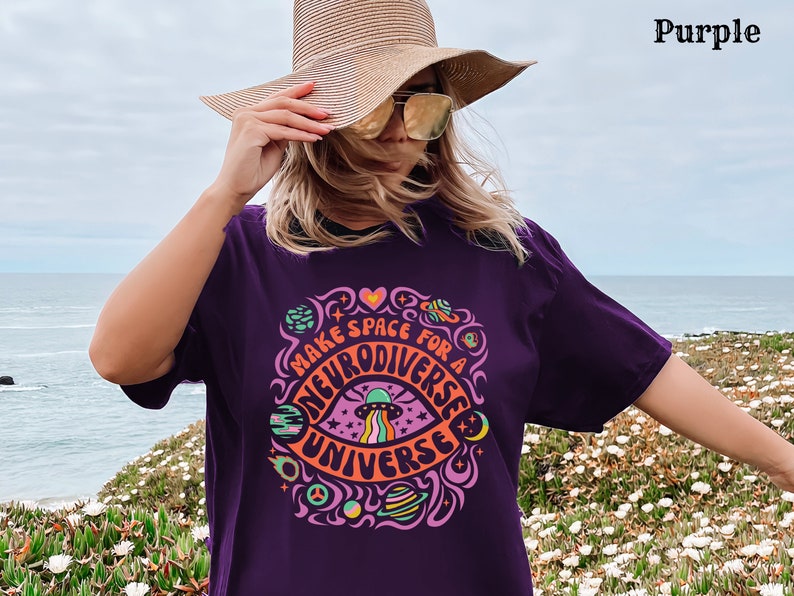 May include: A woman wearing a purple t-shirt with the text "Make Space for a Neurodiverse Universe" in a colourful, psychedelic font. The shirt features a graphic of an eye with a UFO flying over it. The woman is standing in front of a body of water and a field of flowers.
