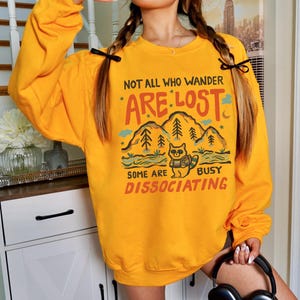 May include: Yellow sweatshirt with a graphic of a raccoon in a forest with the text "Not all who wander are lost. Some are busy dissociating."