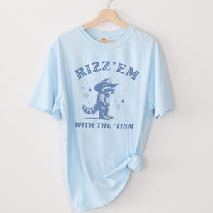 Peut inclure: T-shirt bleu clair avec un graphique bleu d'un raton laveur portant un chapeau de cowboy et le texte "Rizz 'Em With The 'Tism".