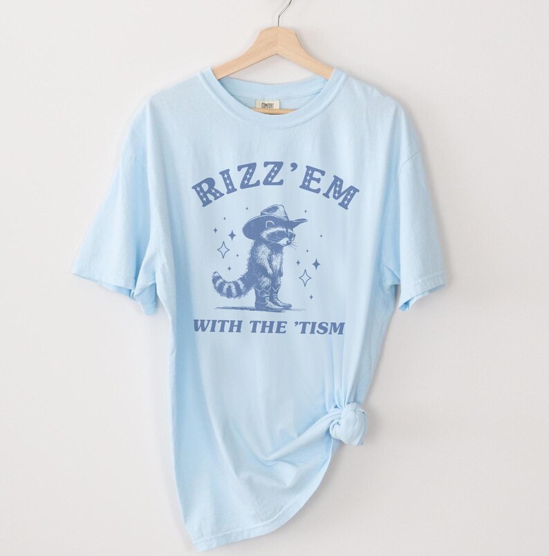 Peut inclure: T-shirt bleu clair avec un graphique bleu d'un raton laveur portant un chapeau de cowboy et le texte "Rizz 'Em With The 'Tism".