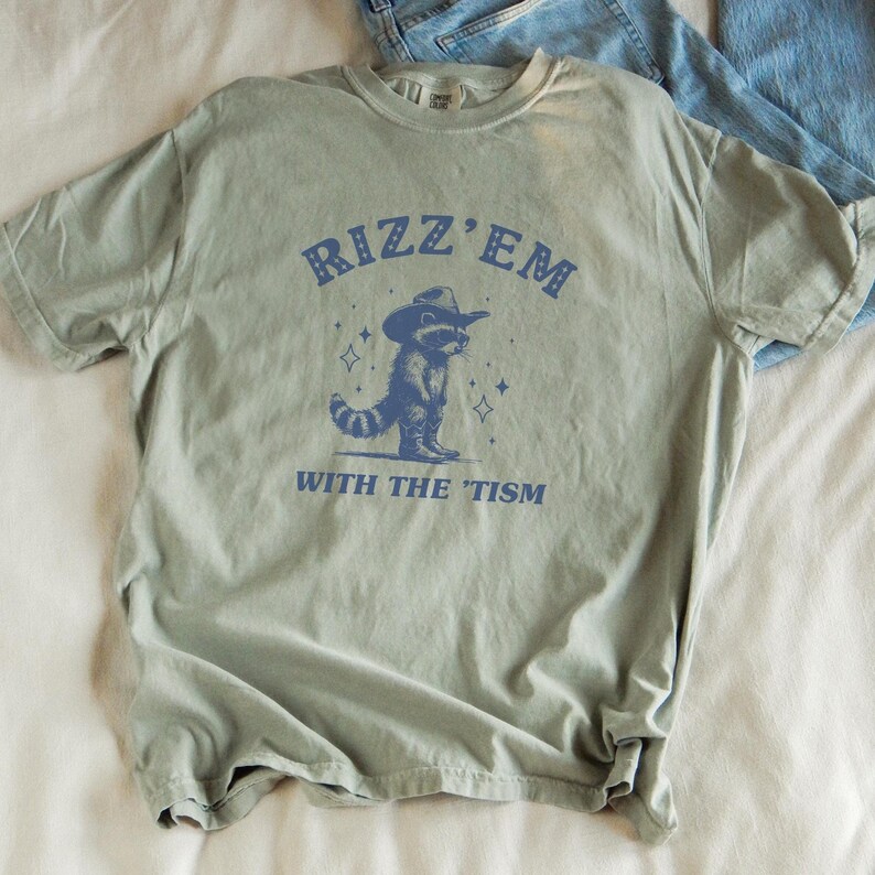 Peut inclure: Un t-shirt vert avec un raton laveur bleu portant un chapeau de cowboy et le texte "Rizz 'Em With The 'Tism" imprim&eacute; dessus.