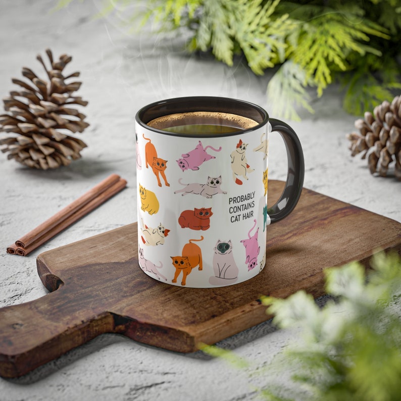 May include: A white ceramic mug with a black handle and a design of colourful cats. The mug has the text "PROBABLY CONTAINS CAT HAIR" printed on it.