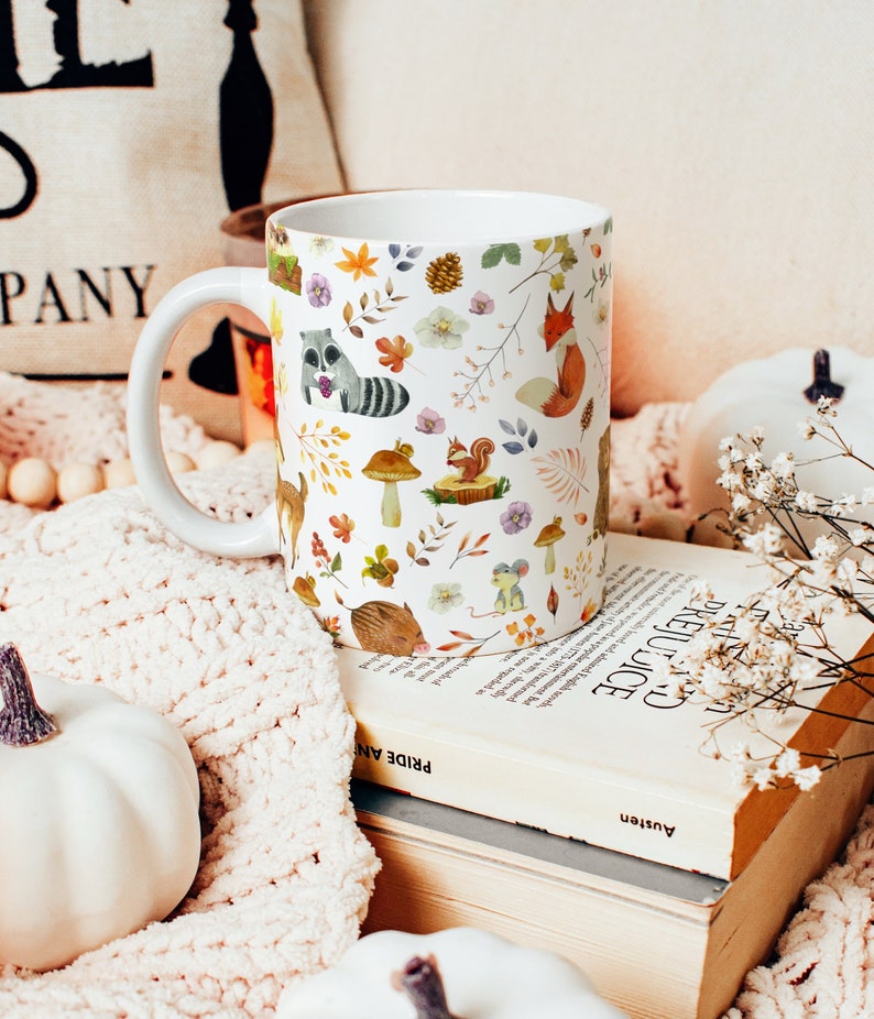 May include: White ceramic mug with a autumn-themed design featuring woodland animals, leaves, and mushrooms. The mug has a large handle and is sitting on a stack of books.