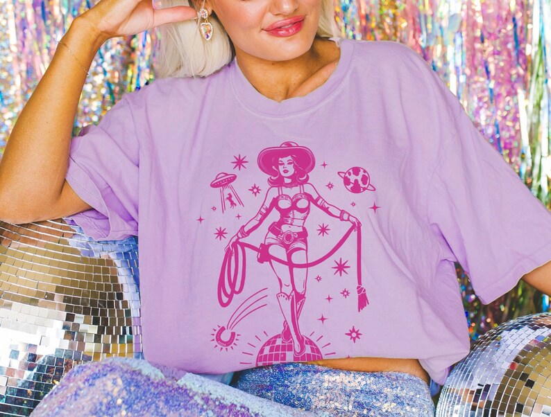 May include: A light purple t-shirt featuring a pink graphic of a cowgirl with a lasso, standing on a disco ball, with a UFO and planet in the background. The shirt has short sleeves and a relaxed fit.