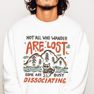 May include: White sweatshirt with a graphic print of a raccoon in a forest setting. The text reads "Not all who wander are lost, some are dissociating."