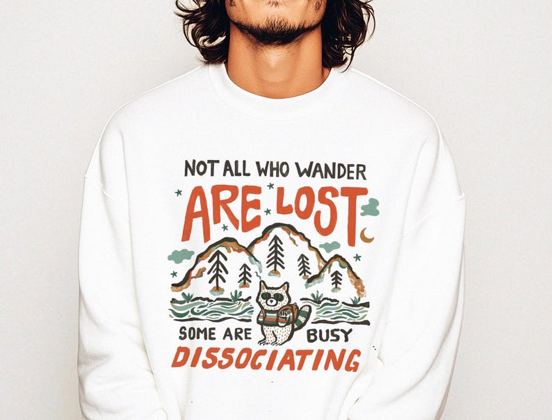 May include: White sweatshirt with a graphic print of a raccoon in a forest setting. The text reads "Not all who wander are lost, some are dissociating."