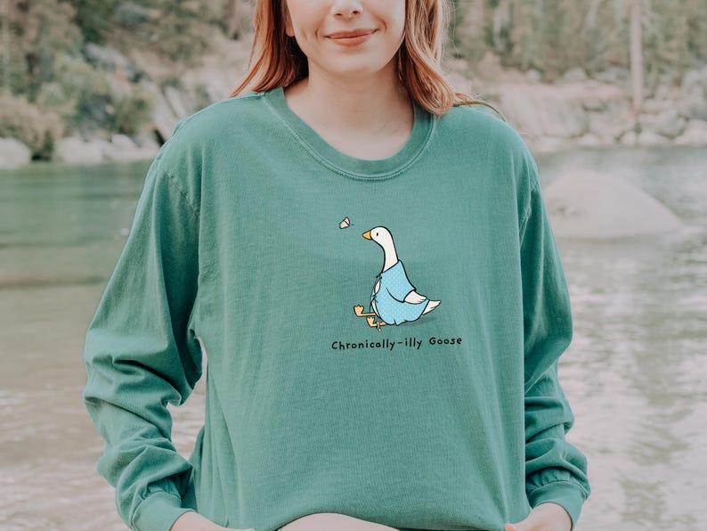 May include: A teal long-sleeve shirt with a cartoon goose wearing a blue jacket and the text "Chronically-Illy Goose" printed on the front.