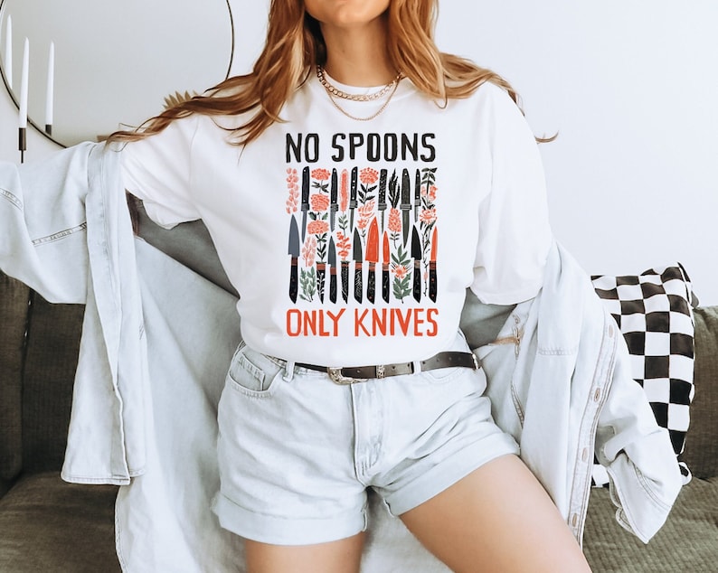 May include: White long-sleeved t-shirt with a graphic of black knives and red flowers. The text "NO SPOONS ONLY KNIVES" is printed on the shirt.