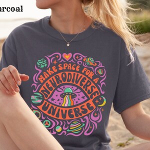 May include: A charcoal grey t-shirt with a colourful graphic design featuring a psychedelic eye with a UFO and the text "Make space for a neurodiverse universe".