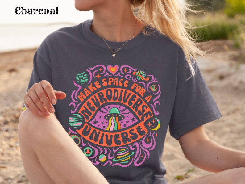 May include: A charcoal grey t-shirt with a colourful graphic design featuring a psychedelic eye with a UFO and the text "Make space for a neurodiverse universe".