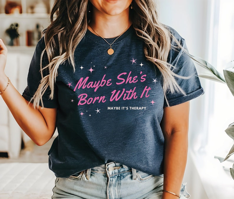 Puede incluir: Una camiseta azul marino con el texto "Maybe She's Born With It" en letras rosas y las palabras "Maybe It's Therapy" en letras rosas m&aacute;s peque&ntilde;as debajo. La camiseta tiene un corte casual y mangas cortas.