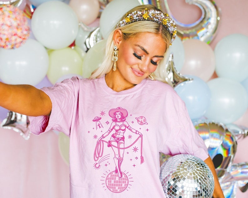 May include: A light pink t-shirt featuring a pink graphic of a cowgirl on a disco ball, with a lasso, stars, and a UFO. The cowgirl wears a hat and boots. The background includes balloons and a disco ball.