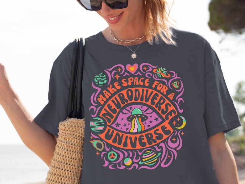 May include: A dark grey t-shirt with a colourful graphic design featuring a psychedelic eye with a UFO inside, surrounded by planets and stars. The text reads "Make space for a neurodiverse universe".