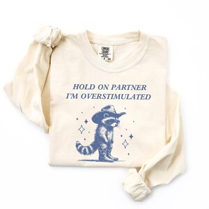 K&ouml;nnte beinhalten: Ein beiges Langarmshirt mit dem Text "HOLD ON PARTNER I'M OVERSTIMULATED" und einem Cartoon-Waschb&auml;ren, der einen Cowboyhut tr&auml;gt.