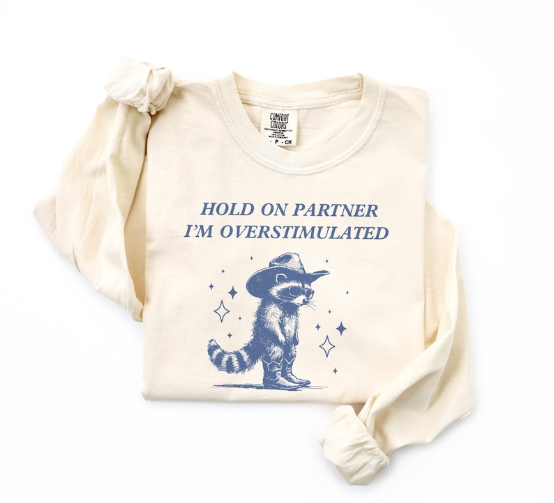 K&ouml;nnte beinhalten: Ein beiges Langarmshirt mit dem Text "HOLD ON PARTNER I'M OVERSTIMULATED" und einem Cartoon-Waschb&auml;ren, der einen Cowboyhut tr&auml;gt.